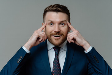 Portrait of handsome male entrepreneur keeps fingers on temples looks with happy surprised expression at camera tries to recall information isolated over grey background. Experienced businessman