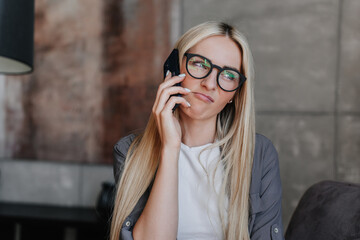 Young Swedish blonde woman in glasses, grey shirt sitting on chair talks with friend hesitating making choice. Attractive caucasian businesswoman annoyed of conversation. Doubt.