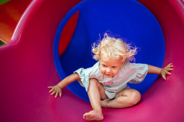 A child of 4 years old curly blonde girl plays on the playground in a purple plastic tube.