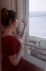 Portrait of woman in red dress from 19th century at home looking through the window curiously to the snowy field