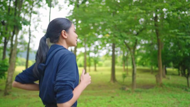 Young asian woman in dark blue hoody running inside the wooden park, natural exercise track, self fitness motivation, endurance training, body conditioning wellness, runner pulse rate breath control
