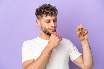 Young caucasian man holding a Bitcoin isolated on purple background and looking up