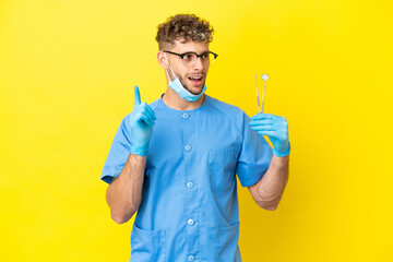 Dentist blonde man holding tools isolated on background thinking an idea pointing the finger up