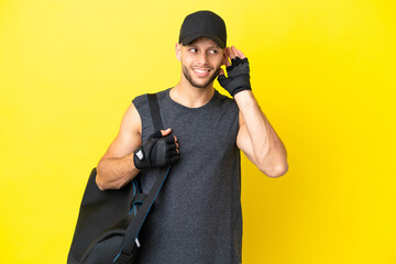 Young sport blonde man with sport bag isolated on yellow background listening to something by...