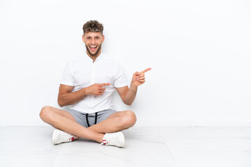 Young blonde man sitting on the floor isolated on white background surprised and pointing side