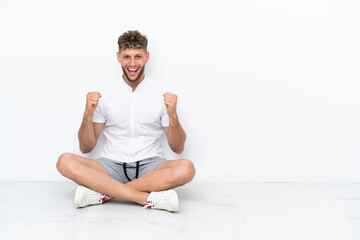 Fototapeta premium Young blonde man sitting on the floor isolated on white background celebrating a victory in winner position