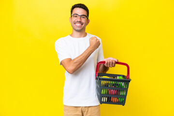 Young handsome man holding a shopping basket full of food over isolated yellow background...