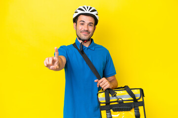 Young handsome man with thermal backpack over isolated yellow background showing and lifting a finger