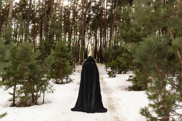 Blonde woman in a black cloak with a hood in winter in a pine forest. Female in cloak with hood from the view from the back side