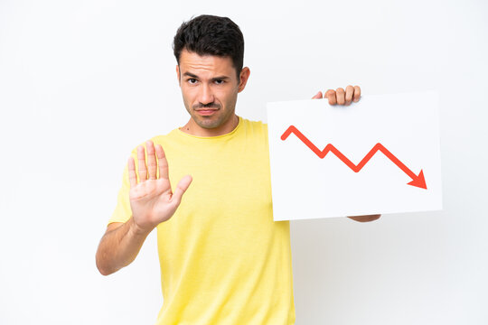 Young handsome man over isolated white background holding a sign with a decreasing statistics arrow symbol and doing stop sign