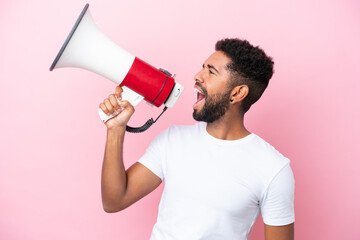 Young Brazilian man isolated on pink background shouting through a megaphone to announce something...