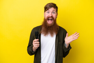 Redhead man with beard picking up a microphone isolated on yellow background with shocked facial expression