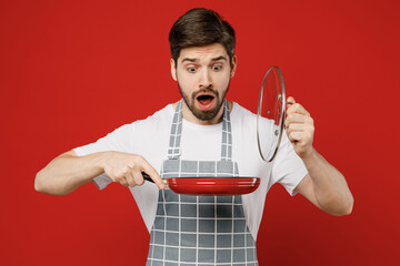 Young confused displeased sad shocked male housewife housekeeper chef cook baker man wearing grey apron hold in hand frying pan open lid isolated on plain red background studio. Cooking food concept.