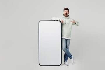 Full body young caucasian man wear mint hoody point index finger on big huge blank screen mobile cell phone smartphone with workspace copy space mockup area isolated on plain solid white background.