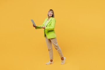 Full body elderly fun IT woman 50s year old wear green jacket white t-shirt hold use work on laptop pc computer look aside isolated on plain yellow background studio portrait People lifestyle concept