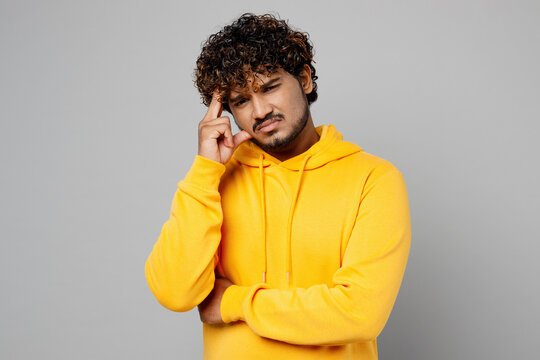 Young minded worried toughtful concerned Indian man 20s he wearing casual yellow hoody looking camera prop up chin think isolated on plain grey background studio portrait. People lifestyle portrait.