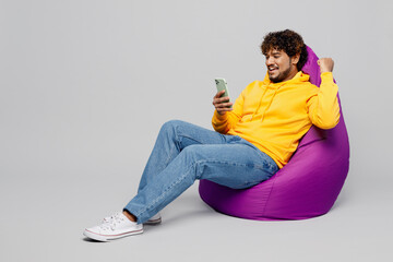 Full body young Indian man wear casual yellow hoody sit in bag chair hold in hand use mobile cell...