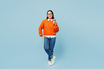 Full body young smiling happy woman of Asian ethnicity wear orange sweater glasses hold takeaway delivery craft paper brown cup coffee to go isolated on plain pastel light blue cyan background studio.