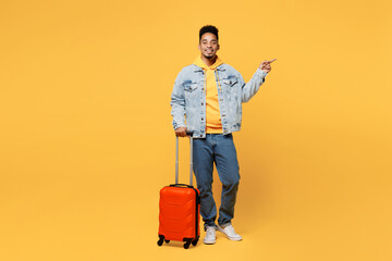 Full body traveler man of African American ethnicity wear casual clothes hold valise point aside isolated on plain yellow background. Tourist travel abroad in free time rest. Air flight trip concept.