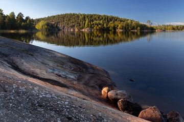 A rocky slope at lake coast and island with dense trees reflected in lake water, Karelia, Russia