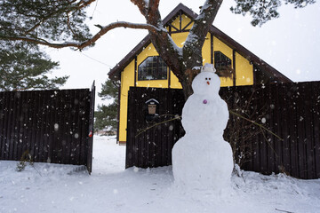 Naklejka premium Funny snowman in a hat and scarf on the background of a yellow house in the yard. Winter, winter entertainment, snowfall