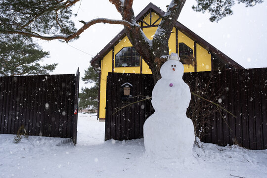 Funny snowman in a hat and scarf on the background of a yellow house in the yard. Winter, winter entertainment, snowfall