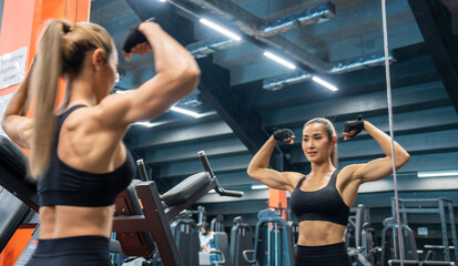 Obraz na płótnie Canvas Asian woman in workout gloves lifting dumbbells looking in mirror looking muscular and building muscles, looking strong and powerful arms in the gym.Training, athlete, workout, exercises concept