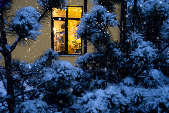 Cozy Festive Window Of The House Outside With The Warm Light Of Fairy Lights Garlands Inside - Celebrate Christmas And New Year In A Warm Home. Christmas Tree, Bokeh, Snow On Pine Trees And Snowfall