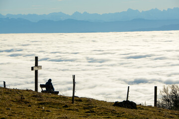 Person resting on bench above a see of fog with panoramic view of the swiss alps and a wooden peak-cross in the foreground from Röti peak in Solothurner Jura, Switzerland