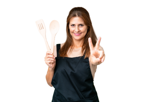 Middle age caucasian woman holding a rolling pin over isolated background smiling and showing victory sign