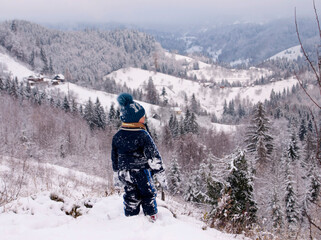 A little boy in a dark blue suit and a blue hat in the snow on a hilltop in winter in the Ukrainian Carpathians, looking at the valley below