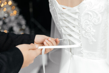 The husband helps the slender bride lace up her wedding white dress on a delicate lace dress. The morning preparation for the wedding describes the newlyweds in detail. Wedding day moments, clothes.