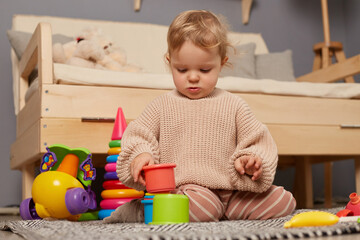 Portrait of little charming baby girl wearing beige sweater playing with colorful plastic toys, sitting on the floor, having fun and. Early learning. Development toys.