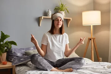 Fotobehang indoor shot of relaxed woman wearing sleeping mask sitting on bed in cozy bedroom, having morning yoga practice, sits in lotus pose with closed eyes and peaceful expression. © sementsova321