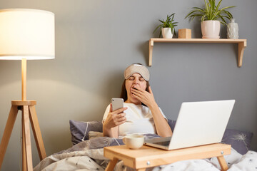 Indoor shot of sleepy tired woman wearing sleep mask sitting with laptop in bed at home, holding mobile phone in hand, looking at screen, yawning and covering mouth with hand.