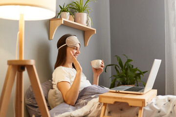 Indoor shot of attractive cheerful woman wearing sleep mask sitting with laptop in bed at home and...