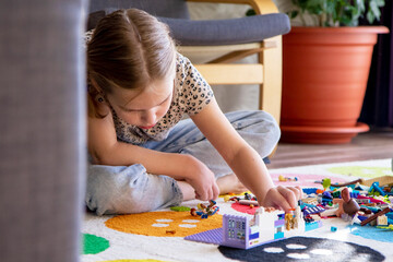 Little blonde girl plays with colorful toy blocks at home sitting on the floor in the living room. Builds and stacks. Educational games for children.