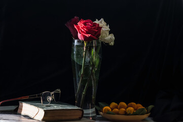 Still life with a closed book and glasses. A glass vase with red and white roses stands on a table covered with a black tablecloth. Ripe oranges with green leaves in a plate.