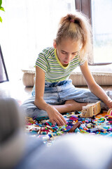 A fair-haired schoolgirl girl sits on the floor in the living room and assembles a designer from multi-colored plastic blocks. Leisure and entertainment after school