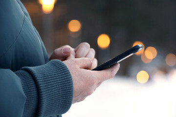 Female hands with smartphone close up on blurred lights background. Woman using mobile phone stands on a street in snow winter