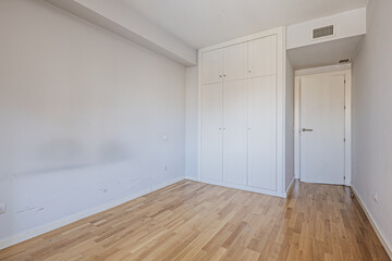 Empty room with blue walls, a three-section built-in wardrobe with mezzanines with smooth white doors, air conditioning through ducts in the ceiling and oak wood floors.