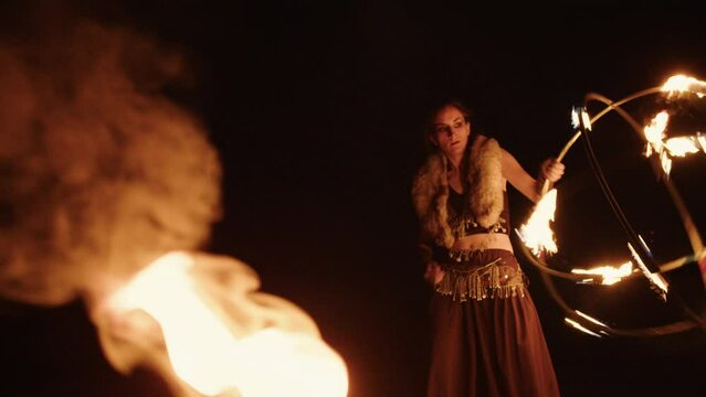 Girl in tribal fusion costume is standing near bonfire, holding, spinning burning sphere around her body. Female firedancer performs at night, revolving flaming round metal rod props at fire show.