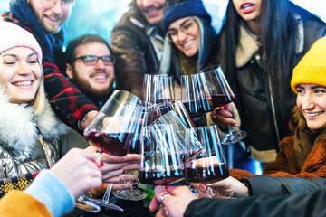 Happy multiracial friends toasting red wine at restaurant patio - Group of young people wearing winter clothes having fun at outdoors winebar terrace - Dining life style and friendship concept 