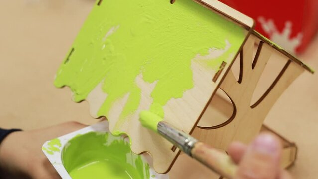 A child's hand paints with the brush the roof of a plywood craft with bright green paint while holding a paint cup. Drawing a homemade birdhouse