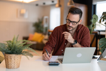Serious millennial man using laptop sitting at the table in a home office. Focused guy in casual clothing looking at the paper, writing down ideas or a future business plan and using laptop.