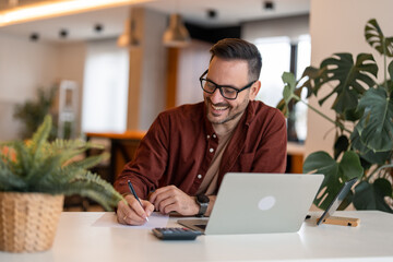 Shot of a young satisfied businessman using a laptop and writing business plan on a paper at his...