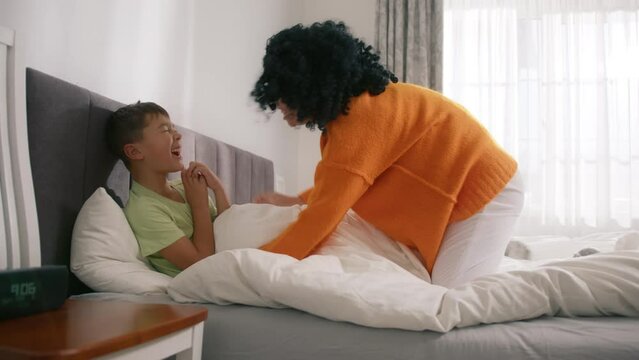 Playing smiling mum enjoying bonding cuddling hugging spending time with funny kid at home. Happy carefree mother and cute small son kid tickling, laughing, having fun lying on bed on weekend morning