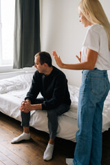 Vertical shot of despaired young husband sitting on bed looking away ignoring angry blonde wife...