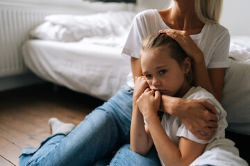 Portrait of sad little girl looking at camera and loving caring mother comforting offended afraid child daughter, showing love and care, expressing support, hugging and stroking hair sitting on floor. - Powered by Adobe