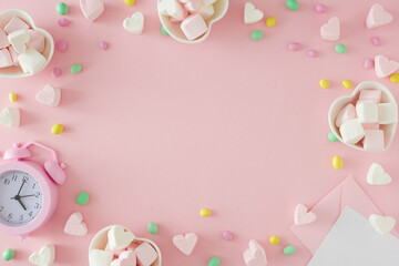 Sweet Valentines day concept. Flat lay photo of heart shaped saucers with candies and heart marshmallow, paper envelope, alarm clock on pastel pink background with copy space in the middle.